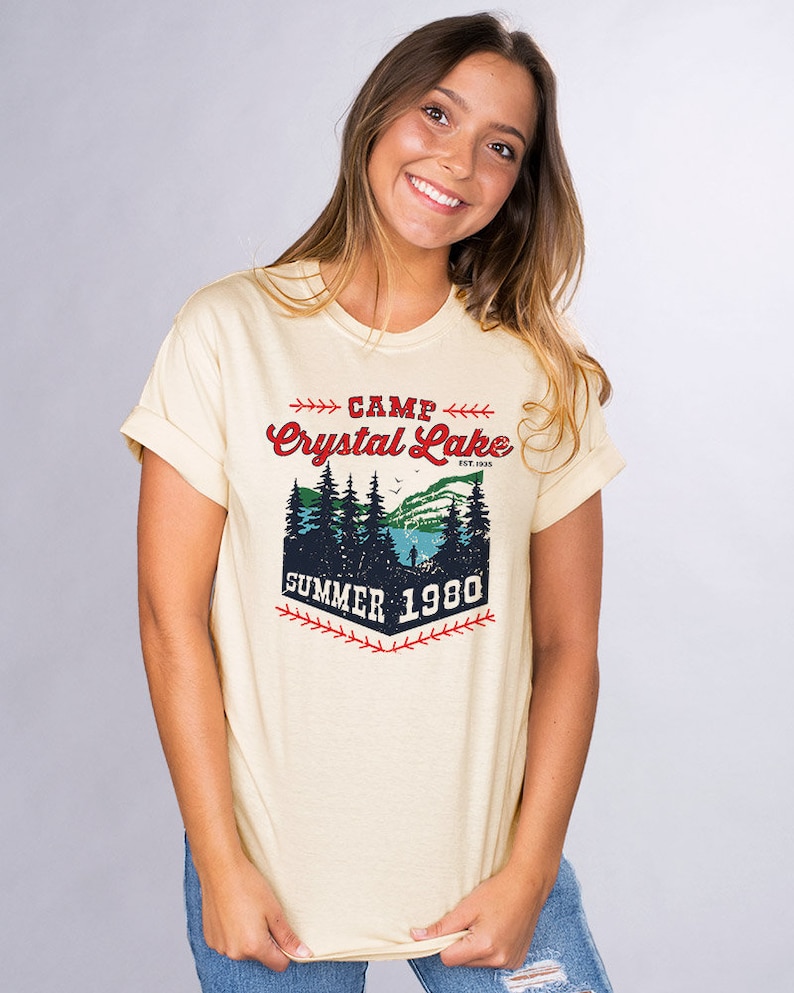 May include: A light yellow t-shirt with a vintage-style graphic design. The design features a forest scene with a lake, trees, and a person fishing. The text reads "Camp Crystal Lake Est. 1925 Summer 1980".