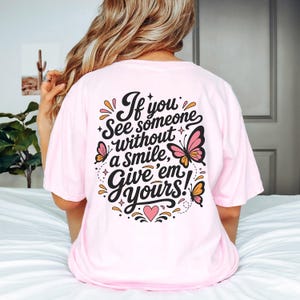 May include: A light pink t-shirt featuring black cursive text on the back that reads, If you see someone without a smile, give 'em yours! The design includes three colorful butterflies in shades of pink and orange, small stars, and a pink heart.