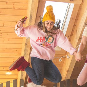 May include: A woman wearing a pink sweatshirt with the text "HAVE A GOOD DAY" in colourful letters, a yellow beanie, and blue jeans jumps in the air while holding a glass of champagne. She is holding hands with another person out of frame.