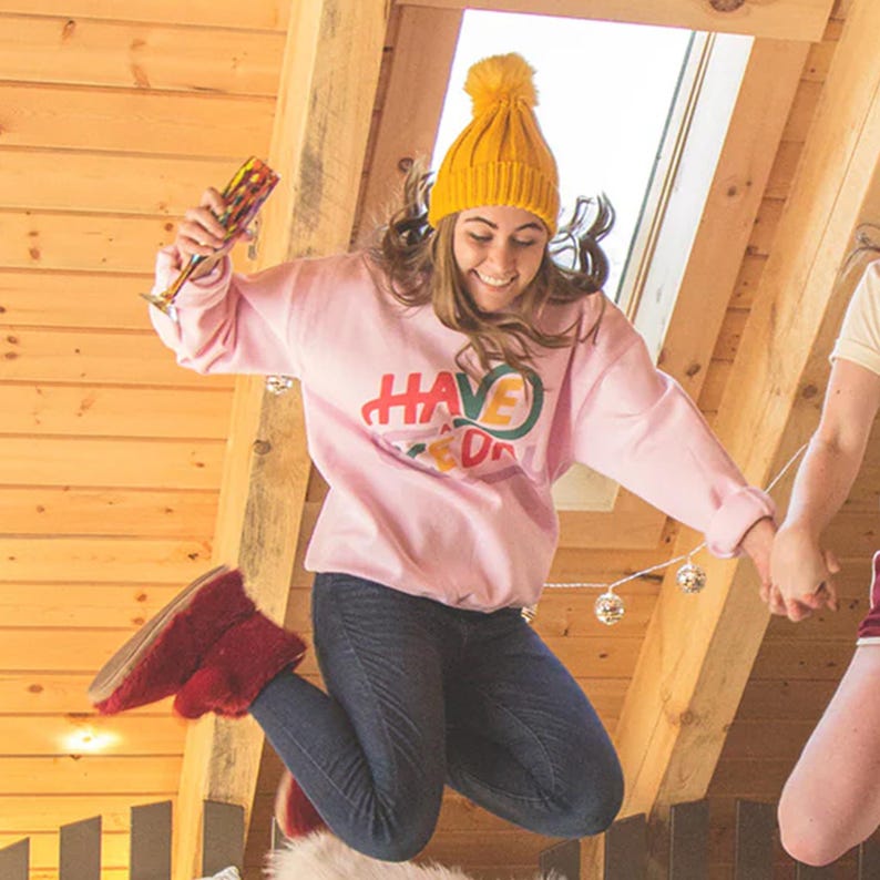 May include: A woman wearing a pink sweatshirt with the text "HAVE A GOOD DAY" in colourful letters, a yellow beanie, and blue jeans jumps in the air while holding a glass of champagne. She is holding hands with another person out of frame.