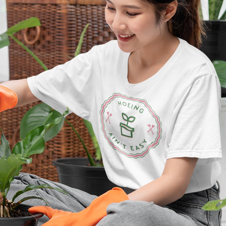May include: A white t-shirt with a green and pink circular graphic that says "Hoeing Ain't Easy" with a small plant in a pot inside the circle.