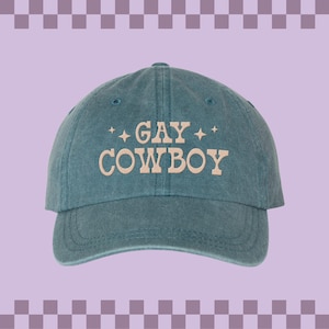 May include: A teal blue baseball cap with the text "GAY COWBOY" embroidered in beige on the front.