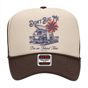May include: A brown and beige trucker hat with a vintage illustration of a van with surfboards on top. The text "Don't Bug Me I'm on Island Time Surf Club" is printed on the front of the hat.