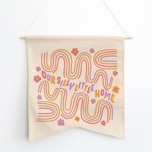 May include: A beige canvas wall tapestry with a colorful abstract design featuring wavy lines in orange, pink, and purple. The text "Our Silly Little Home" is printed in pink and orange.