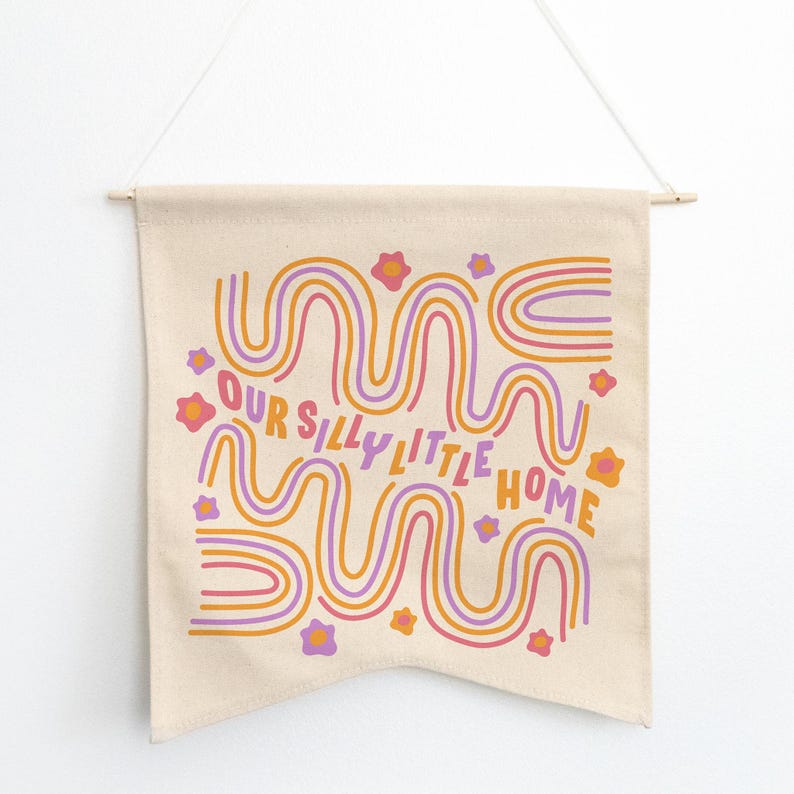 May include: A beige canvas wall tapestry with a colorful abstract design featuring wavy lines in orange, pink, and purple. The text "Our Silly Little Home" is printed in pink and orange.