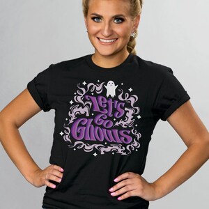 May include: Black t-shirt with the phrase "Let's Go Ghouls" in purple and white lettering. The design includes a ghost and decorative swirls and stars, perfect for Halloween or spooky season.