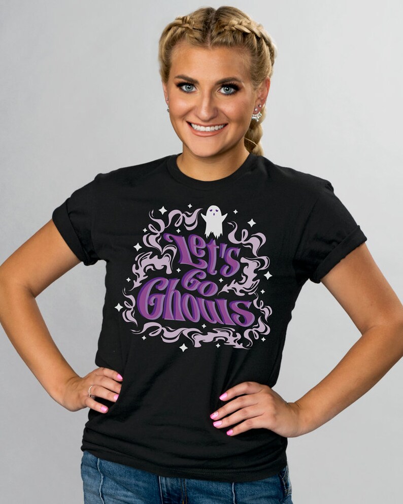 May include: Black t-shirt with the phrase "Let's Go Ghouls" in purple and white lettering. The design includes a ghost and decorative swirls and stars, perfect for Halloween or spooky season.
