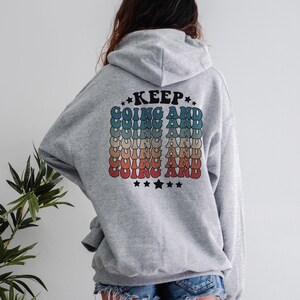 Puede incluir: Sudadera gris con capucha con el texto "KEEP GOING AND" repetido en un patr&oacute;n de arco&iacute;ris en la espalda.