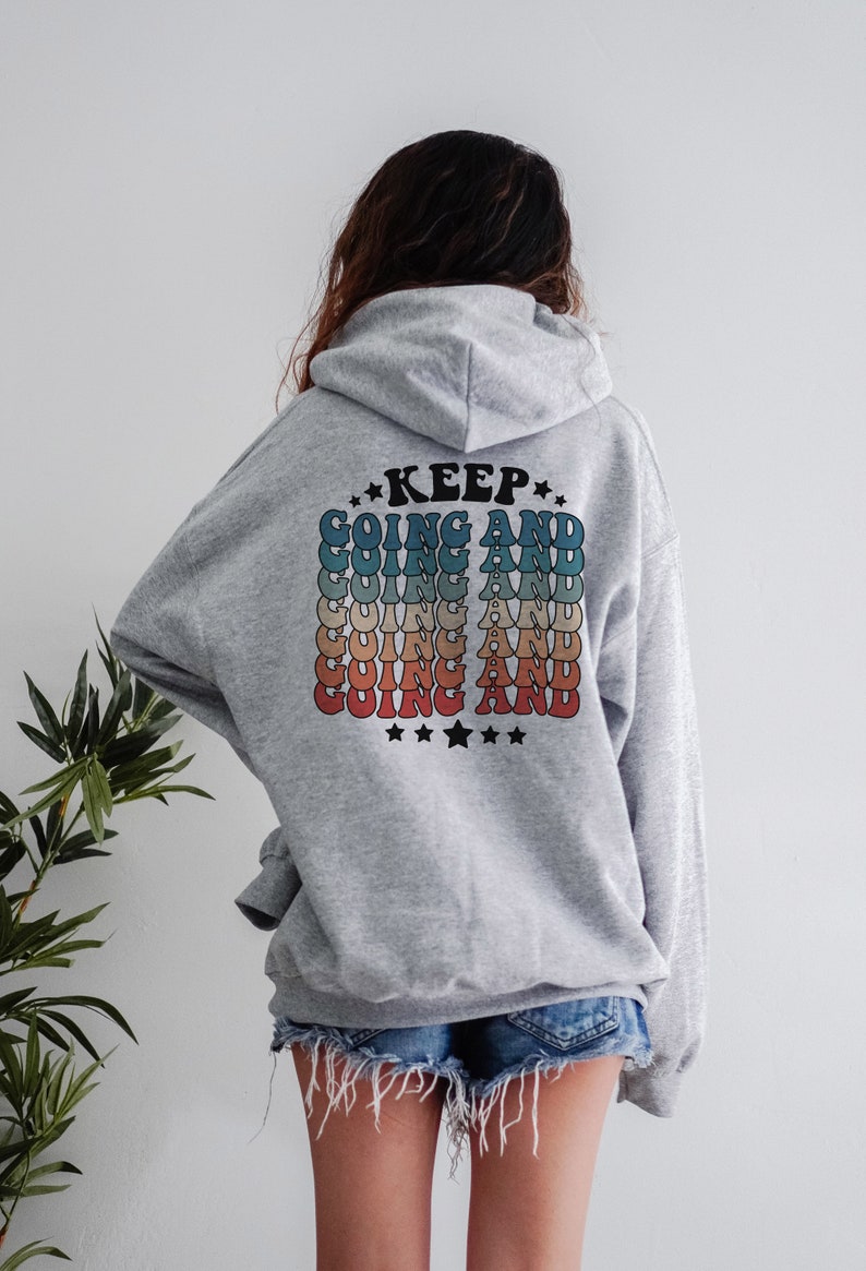 Puede incluir: Sudadera gris con capucha con el texto "KEEP GOING AND" repetido en un patr&oacute;n de arco&iacute;ris en la espalda.