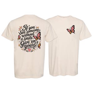 May include: A cream-colored t-shirt featuring a small pink and black butterfly graphic on the front left chest. The back displays the text "If you see someone without a smile, Give 'em yours!" in black script, surrounded by butterflies and a heart.