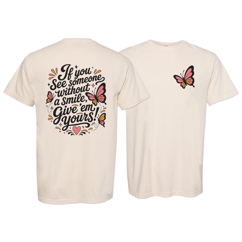 May include: A cream-colored t-shirt featuring a small pink and black butterfly graphic on the front left chest. The back displays the text "If you see someone without a smile, Give 'em yours!" in black script, surrounded by butterflies and a heart.