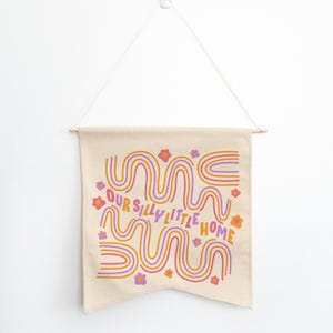 May include: A beige canvas wall hanging with a pink, orange, and purple wavy design and the text "Our Silly Little Home".