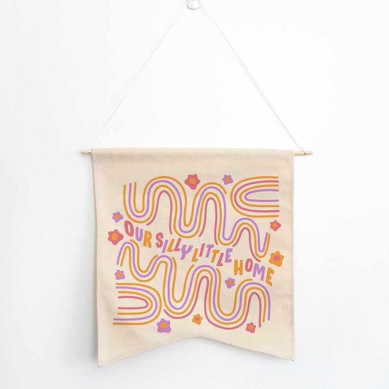 May include: A beige canvas wall hanging with a pink, orange, and purple wavy design and the text "Our Silly Little Home".