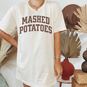 May include: Cream-colored t-shirt with the words "MASHED POTATOES" printed in brown, block letters. The shirt has short sleeves and a relaxed fit. The image is of a person wearing the shirt.