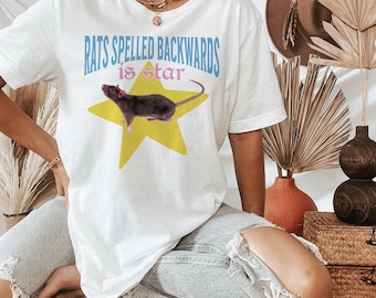 May include: White t-shirt featuring the text "RATS SPELLED BACKWARDS is STAR" in blue and pink. A dark gray rat is centered on a yellow star graphic. The shirt is a casual, unisex style.