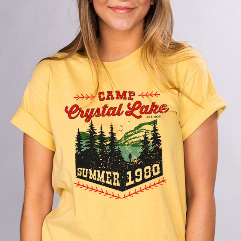 May include: Yellow short-sleeved t-shirt with a vintage-style graphic design. The design features a forest scene with a lake, trees, and the text "Camp Crystal Lake Est. 1935 Summer 1980".