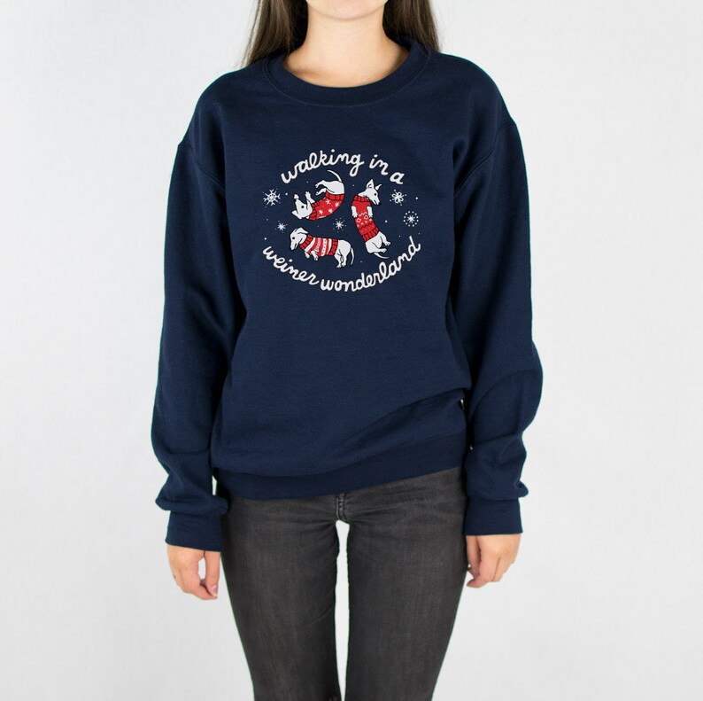 May include: Navy blue sweatshirt with a circular graphic featuring three dachshunds in red and white sweaters, surrounded by snowflakes. The text "walking in a weiner wonderland" is arched around the dogs. The sweatshirt is worn by a person.