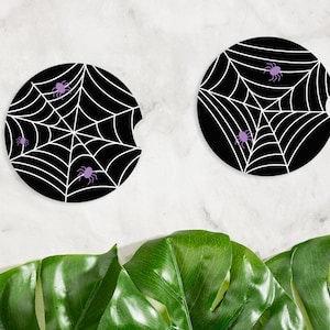 May include: Two black car coasters with white spiderwebs and purple spiders. The coasters have a circular shape with a small notch on one side. The background is a white marble surface with green leaves.