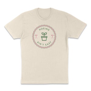 May include: A beige t-shirt featuring a green and pink graphic that reads "HOEING AIN'T EASY" with a plant in a pot and two crossed shovels.