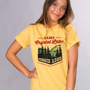 May include: Yellow t-shirt with a vintage-style graphic that says "Camp Crystal Lake Est. 1935 Summer 1980". The graphic features a lake scene with trees and a baseball bat.