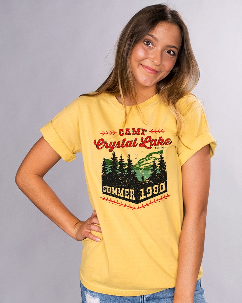 May include: Yellow t-shirt with a vintage-style graphic that says "Camp Crystal Lake Est. 1935 Summer 1980". The graphic features a lake scene with trees and a baseball bat.