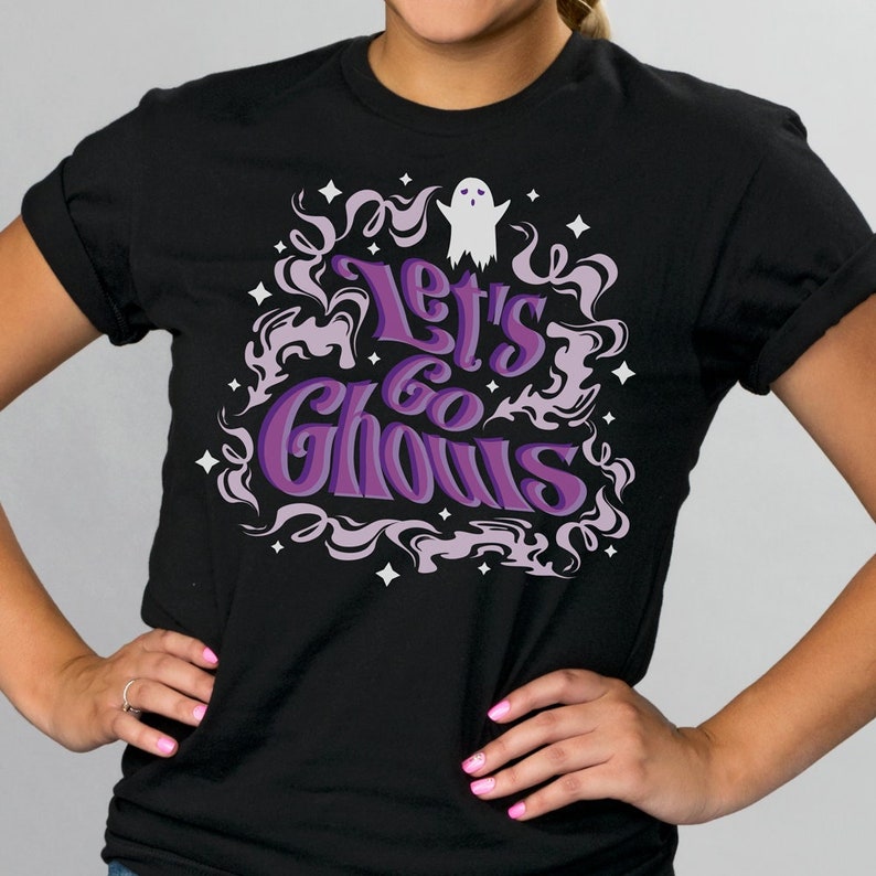 May include: Black t-shirt with the phrase "Let's Go Ghouls" in purple and white, surrounded by a decorative design of swirling smoke and stars. A small white ghost graphic is above the text. The shirt is a crew neck.