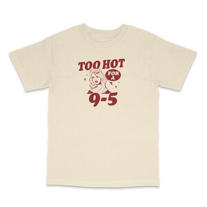 May include: A beige t-shirt with a red graphic of a woman with the text "Too Hot For A 9-5"