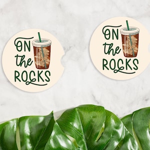 May include: Two round beige coasters with the text "On the Rocks" in green lettering and a cartoon image of a glass of iced coffee.