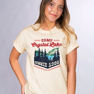 May include: A light yellow t-shirt with a vintage-style graphic design. The graphic features a forest scene with a lake, trees, and a person fishing. The text reads "Camp Crystal Lake Est. 1935 Summer 1980".