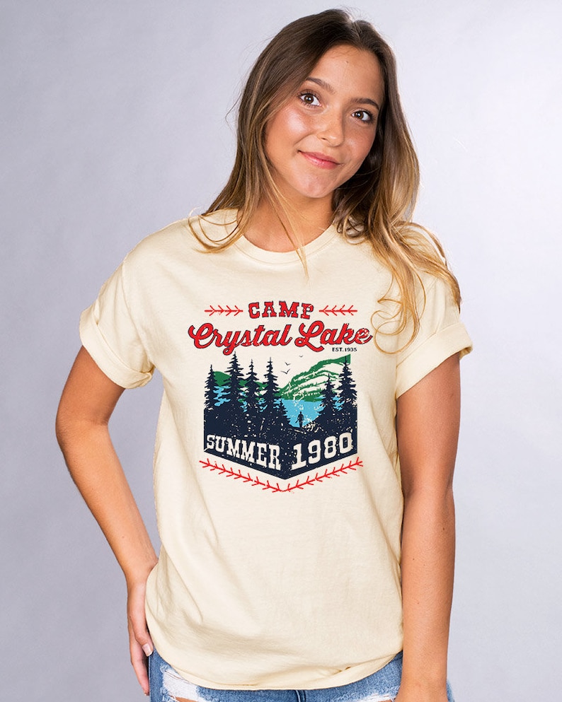 May include: A light yellow t-shirt with a vintage-style graphic design. The graphic features a forest scene with a lake, trees, and a person fishing. The text reads "Camp Crystal Lake Est. 1935 Summer 1980".