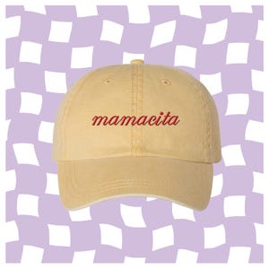 May include: A light yellow baseball cap featuring the word mamacita embroidered in red cursive script across the front panel. The cap has a structured crown, a curved brim, and stitched eyelets on the top panels for ventilation and durability.