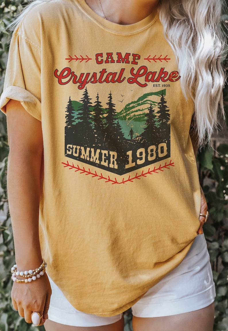 May include: A yellow t-shirt with a vintage-style graphic design that says "Camp Crystal Lake Est. 1935 Summer 1980". The graphic features a forest scene with a lake and a person walking in the distance.
