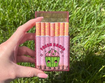 May include: A pink glittery cigarette case with the words "Fresh Baked" and a cartoon cupcake design. The case holds several cigarettes with pink and orange paper. The case is held in a hand against a background of green grass.
