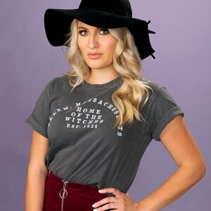 Puede incluir: Una mujer con un sombrero negro de ala ancha, una camiseta gris oscuro con el texto "Salem, Massachusetts, Home of the Witches, Est. 1626", y una falda de pana color burdeos.
