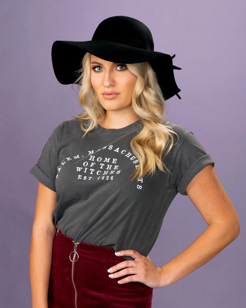 Puede incluir: Una mujer con un sombrero negro de ala ancha, una camiseta gris oscuro con el texto "Salem, Massachusetts, Home of the Witches, Est. 1626", y una falda de pana color burdeos.