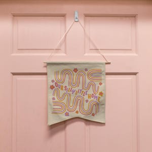 May include: A beige canvas banner with a colorful design of wavy lines and flowers. The banner reads "Our Silly Little Home".