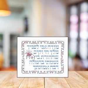 May include: A white framed cross-stitch with a red and blue Hebrew inscription and a small red house with a red roof and a yellow sun. The inscription reads "Blessed is the home that is filled with the light of the Torah".
