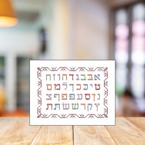 May include: A framed embroidery featuring Hebrew letters in various colors. The letters are arranged in rows and surrounded by a decorative border. The embroidery is displayed on a wooden surface.