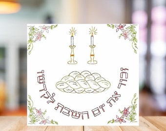 Könnte beinhalten: Eine weiße Karte mit einem grünen und rosafarbenen floralen Rand. In der Mitte der Karte befindet sich ein geflochtenes Challah-Brot, zwei Kerzenleuchter mit brennenden Kerzen und der hebräische Text "Shabbat Shalom" und "L'chaim".