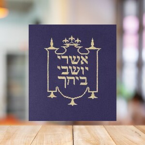 May include: A square, navy blue fabric with gold embroidery. The embroidery features Hebrew text within a decorative frame, topped with a crown. The design is centered on the fabric, creating a symmetrical and elegant appearance.