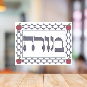 May include: A white cross-stitch embroidery with a black Hebrew word "Shalom" in the center, surrounded by a black and white geometric pattern and red apples at each corner.