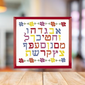 May include: A framed cross-stitch piece with a white background and a colorful Hebrew alphabet. The frame is red and the letters are in different colors, including red, yellow, blue, green, and purple. The letters are surrounded by a border of small flowers.