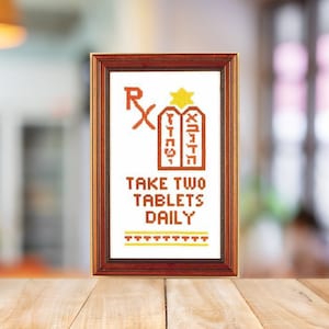 Könnte beinhalten: Ein gerahmtes Kreuzstich-Design mit weißem Hintergrund und brauner und gelber Stickerei. Das Design zeigt zwei Tafeln mit hebräischem Text, einen Stern und den Text "Nehmen Sie zwei Tabletten täglich ein".