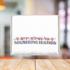 May include: White kitchen towel with a blue and red embroidered design that says "על ידי רחיצת ידיים" in Hebrew and "WASHING HANDS" in English.