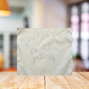 May include: Off-white embroidered cloth with blue stitching. The design features a table setting with a tablecloth, dishes, and text in Hebrew. Decorative swirls and text are embroidered around the table setting.