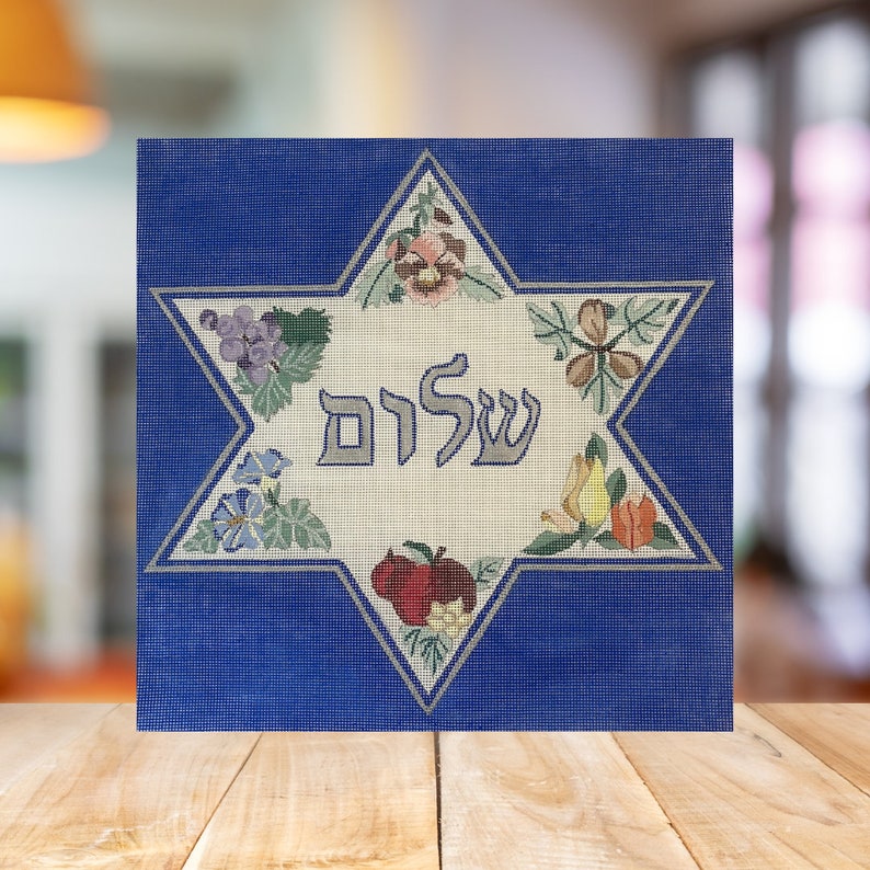 K&ouml;nnte beinhalten: Ein gestickter Davidstern in Blau und Wei&szlig; mit dem hebr&auml;ischen Wort &bdquo;Shalom&ldquo; in der Mitte. Der Stern ist von einer Vielzahl bunter Blumen und Fr&uuml;chte umgeben.
