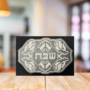 May include: White lace doily with Hebrew text. The doily is rectangular with an ornate, openwork design. The text is centered and appears to be a religious inscription. The doily is displayed on a black background.