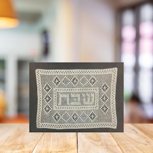 Puede incluir: Un tapete rectangular blanco con un intrincado diseño de encaje. El tapete presenta un diseño rectangular central con texto hebreo, rodeado de motivos florales y geométricos. El tapete está sobre un fondo oscuro.