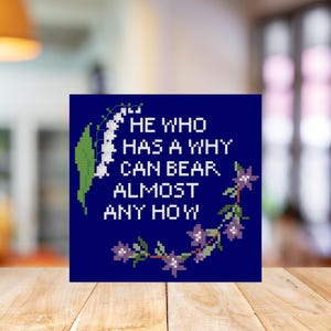 May include: A square, navy blue cross-stitch design with the quote, "HE WHO HAS A WHY CAN BEAR ALMOST ANY HOW" in white. The design includes a floral wreath with purple flowers and green leaves.
