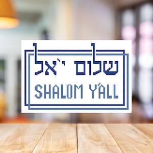 May include: A white rectangular sign with blue text and a blue border. The sign says "Shalom Y'all" in English and Hebrew. The sign is on a wooden surface.